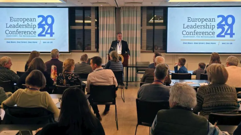 a man standing at a lectern and speaking to people who are seated indoors