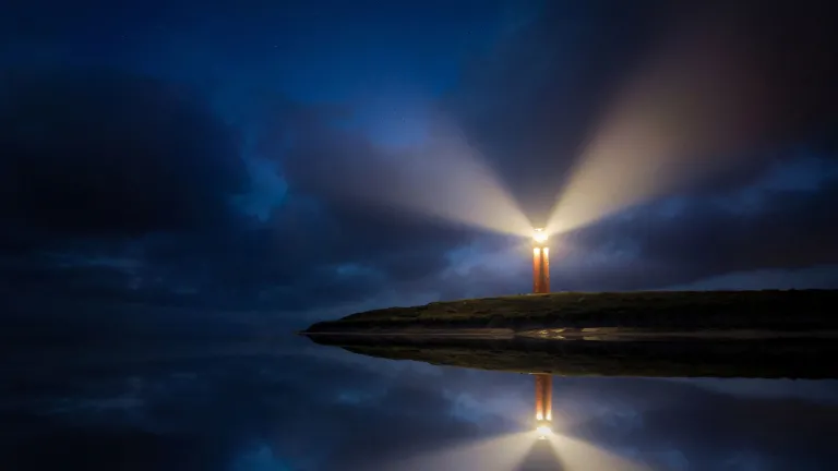 Eierland Lighthouse–Texel, Netherlands a lighthouse sending out two beams of light in the darkness of night and reflecting on the water below