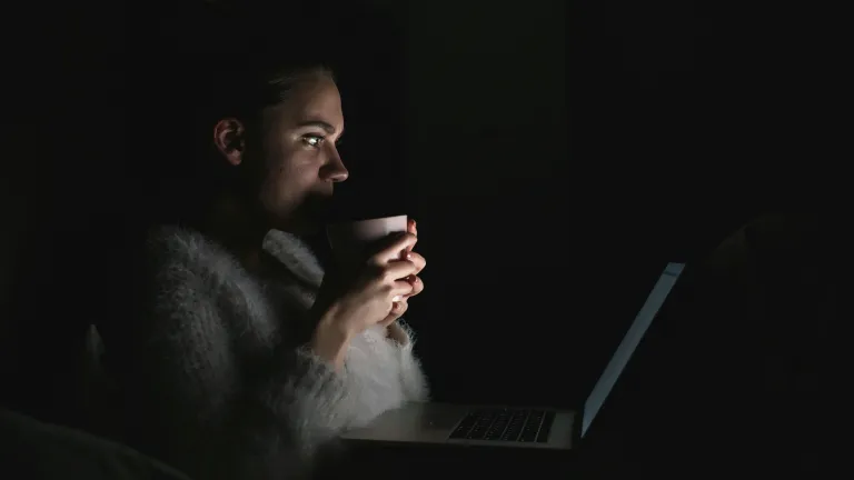a woman looking at the laptop screen in a dark room and holding a coffee mug close to her face