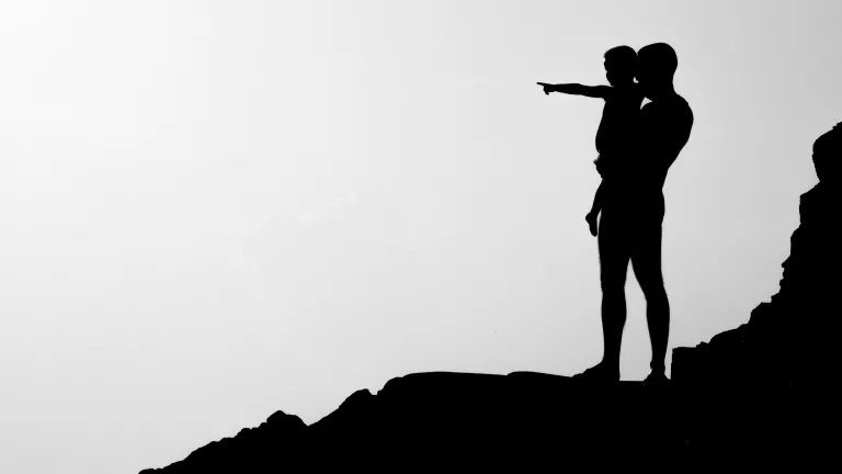 a silhouette of a father holding his son while standing on a rocky surface; the son is pointing into the distance