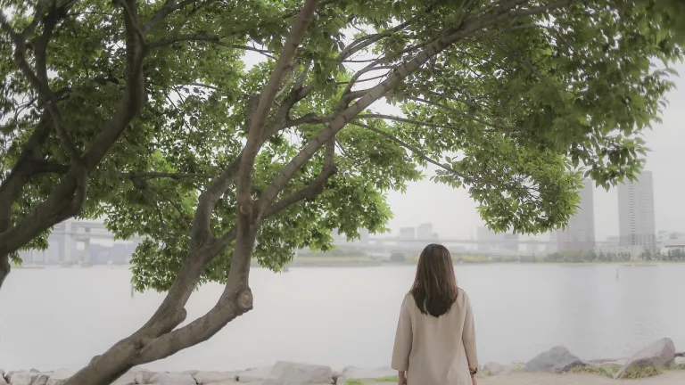 While God created us for companionship, regularly practicing intentional solitude is vital for our spiritual, mental, and emotional well-being—enabling deeper connection with God, greater self-awareness, clearer thinking, and healthier relationships. A woman standing under a tree and looking into the distance at a cityscape.