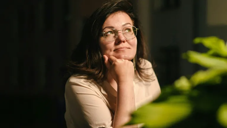 a woman seated behind a plant and looking off into the distance with her chin resting on her hand