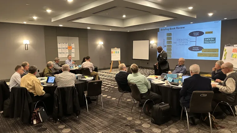 a group of people indoors seated at round tables and listening to a presenter