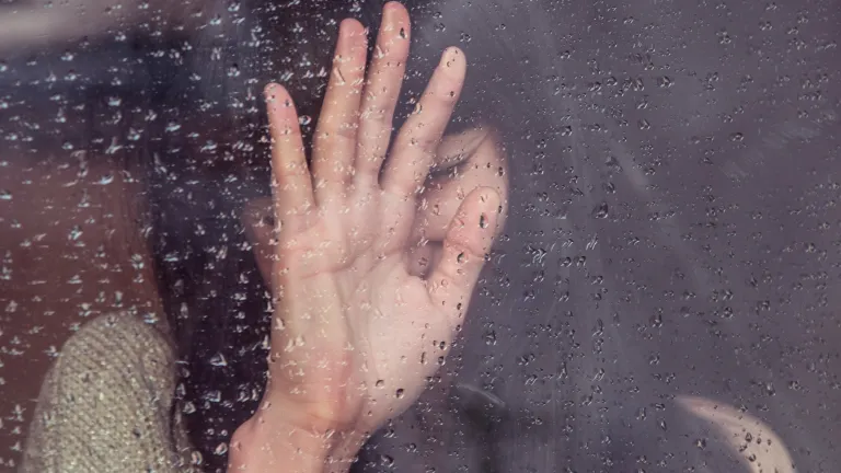 a sad woman with her hand up against a pane of glass dotted with raindrops