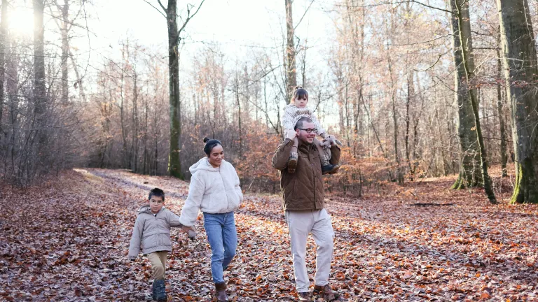 A mom and dad walking in the woods with their two kids. The daughter is on the shoulders of the dad and the son is holding hands with the mom.