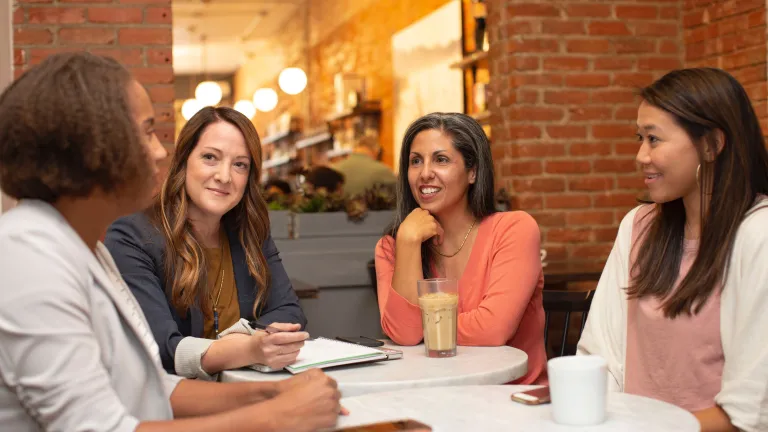 Communication involves more than just talking or writing—it requires listening. a group of four women dressed in business casual sitting at a table in a cafe with a brick wall that has a window looking into the rest of the cafe