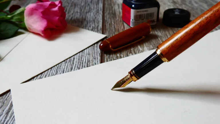 a fountain pen, bottle of ink, blank page, envelope and rose on a table