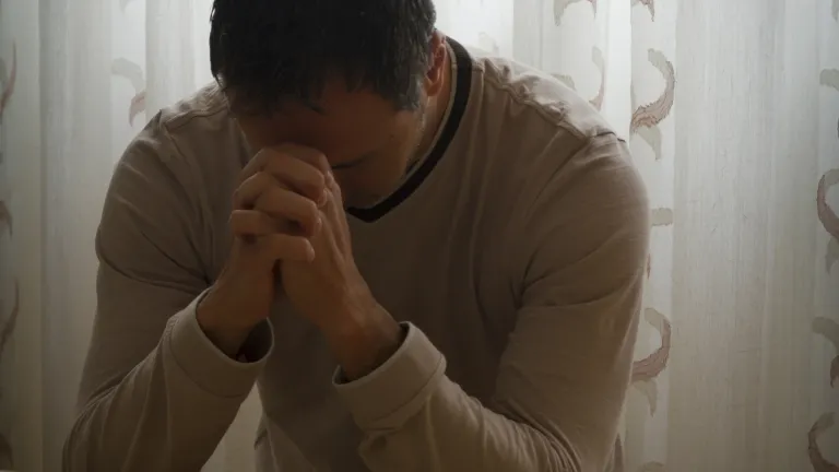 A man praying at the side of a bed.