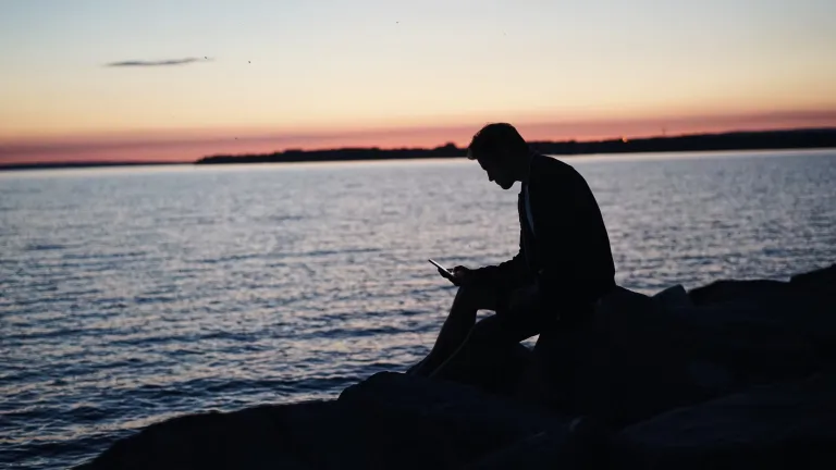 A man sitting on rocks by a body of water looking down at his phone.