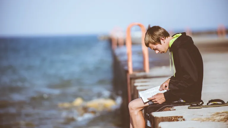A young man reading a Bible while sitting on a dock.