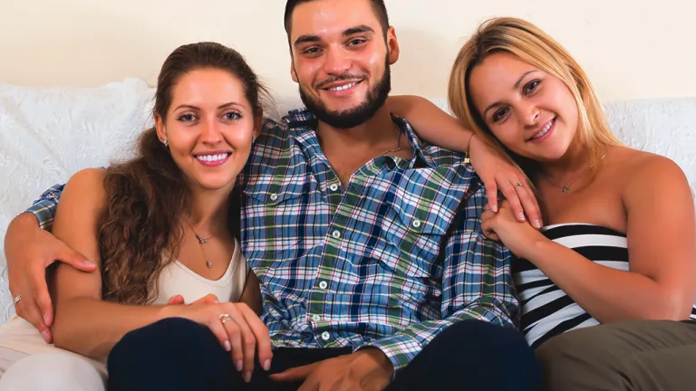 In a society in which nearly any kind of sexual relationship is celebrated, it’s frankly surprising that polygamy remains illegal. A man sitting with two women.