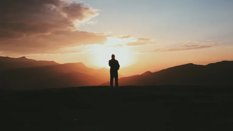 A silhouette of a person stand on a hill with the sun in the background.