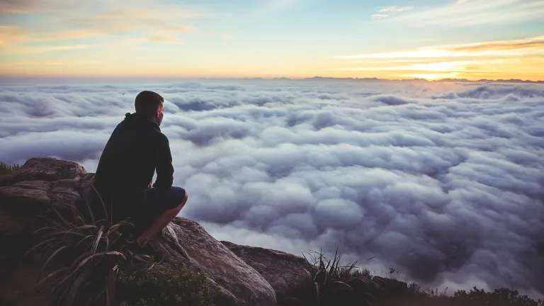 Photo of man thoughtfully squatting at the top of a cliff looking at the clouds.