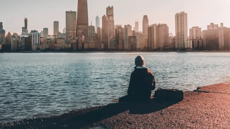Wherever we turn we are being persuaded against a belief in God. A person sitting at the edge of water looking at a city skyline.