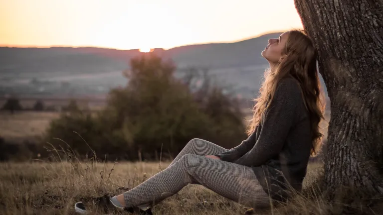 A young woman sitting on the ground leaning against a tree looking up toward the sky.