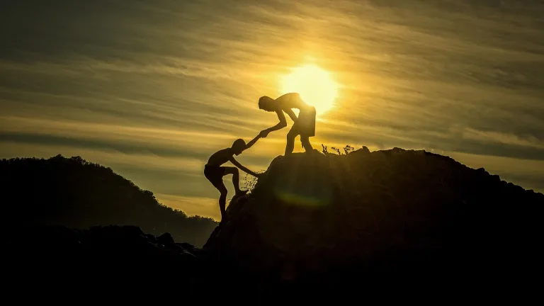 a person helping another climbing up a hill silhouetted against the golden sun in a cloudy sky