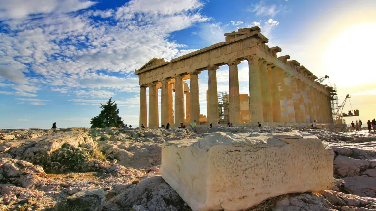 Completed in 438 BC and adorned with fabulous sculptures and statures, the Parthenon stood almost intact until it was severely damaged 1687 by Venetians fighting the Turks. Ancient ruins of the Parthenon with tourists by the structure and a large block of stone in the foreground