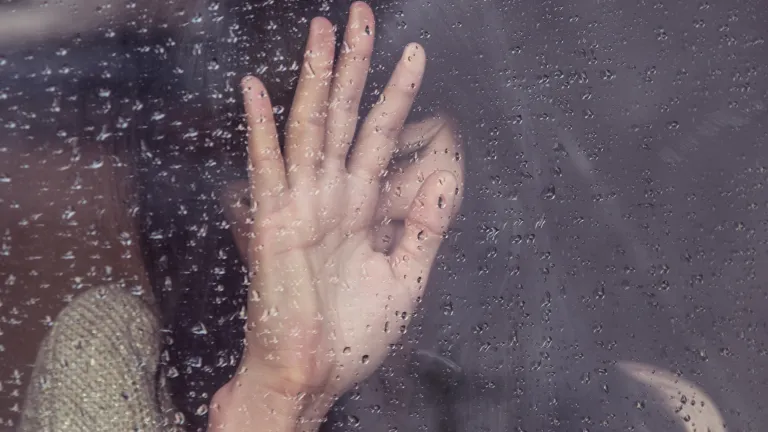 As Christians, we need to open up and allow ourselves to receive the gift of healing for our broken hearts. Woman with her hand up standing behind a window.