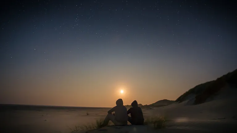 Two people sitting on a beach with the sun setting.