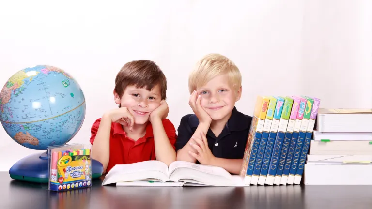 two boys with an open textbook on a desk alongside a globe and other school supplies