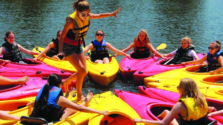 A picture of a group of campers in kayaks form a circle with the kayaks while one camper walks around the circle.