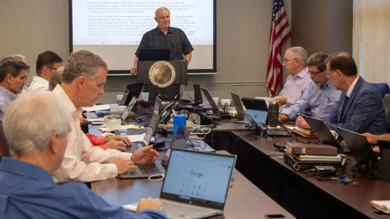 several members of the council of elders seated around a table as one elder presents at the lectern with a slideshow behind him