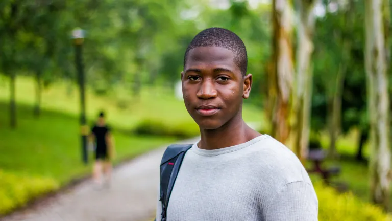 a young man standing outside with a path and green trees in the background and a backpack on one shoulder