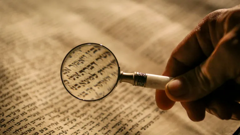 a hand holding a magnifying glass over a scroll of Hebrew text
