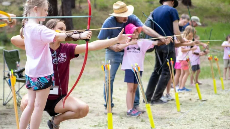 a group of staff assisting preteen girls with archery outdoors