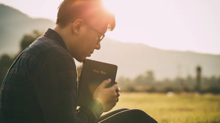 a man sitting outdoors praying while holding Bible