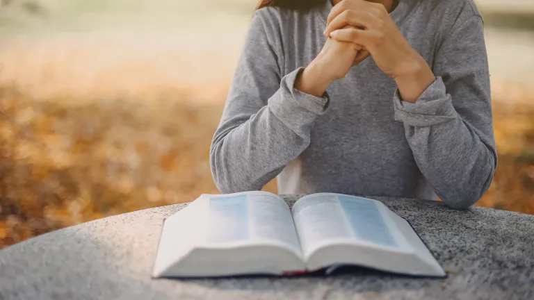 a woman with folded hands sitting with a Bible open in front of her on a table