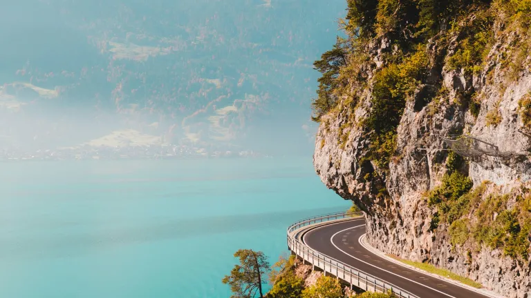a winding road on the side of a cliff above a body of water and blue mountains cape in the distance