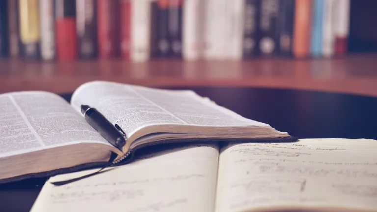 An open Bible and notebook on a table in front of a bookshelf