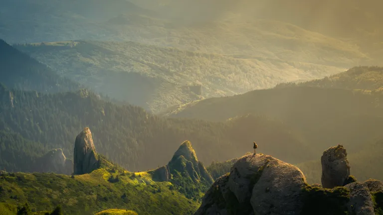 Photo of a person standing atop a mountain, overlooking mountains and the valley at golden hour.