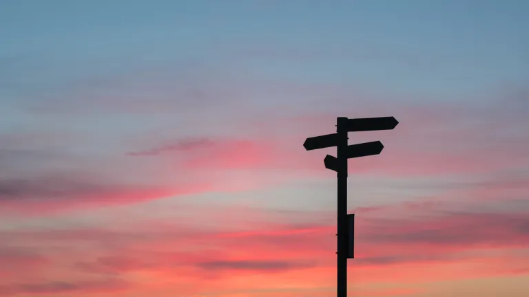 Photo of a signpost with wooden arrows, silhouetted against a pink sunrise sky