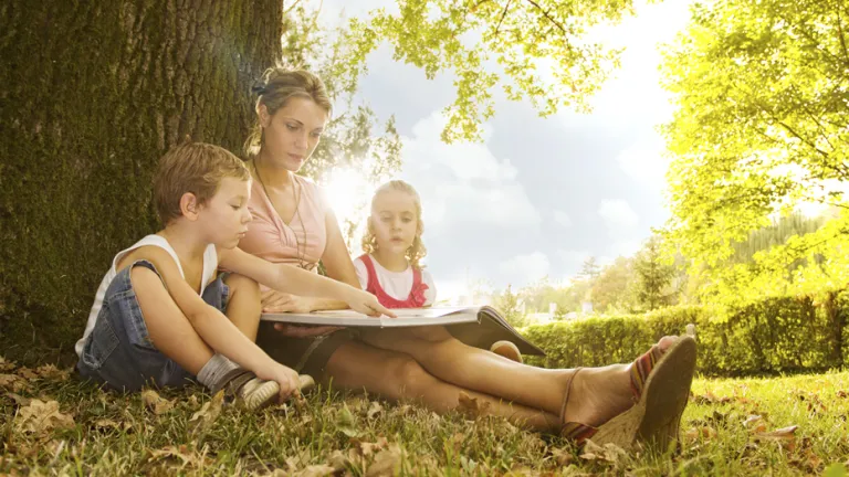 A woman sitting outside beneath a tree reading to her two children.