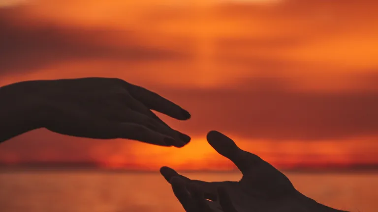 one hand reaching for another silhouetted against a sunset over water