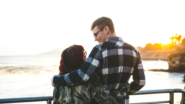 a man with his arm around a woman as they look at each other and stand by a railing overlooking a body of water