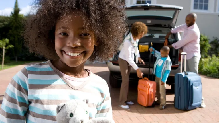 Girl in the forefront smiling with mother, father, and sibling in the background packing up the car with suitcases