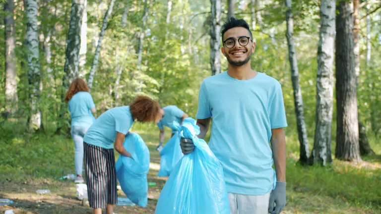a man holding up a plastic trash bag while a team behind him cleans up trash in the forest