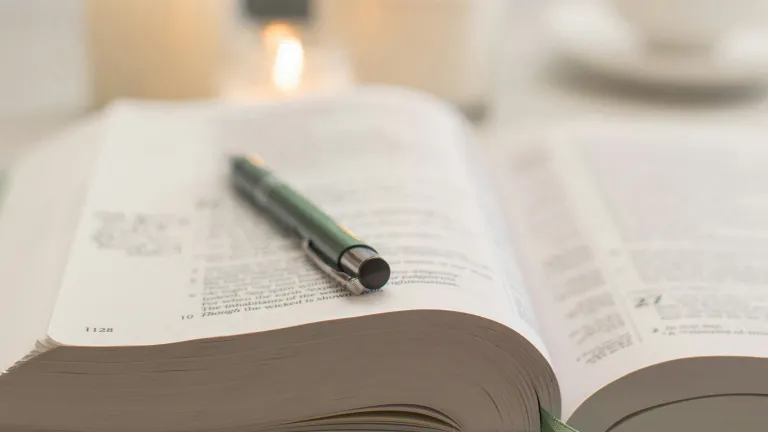 a bible with a pen lying across its open pages and candles and a teacup in the background