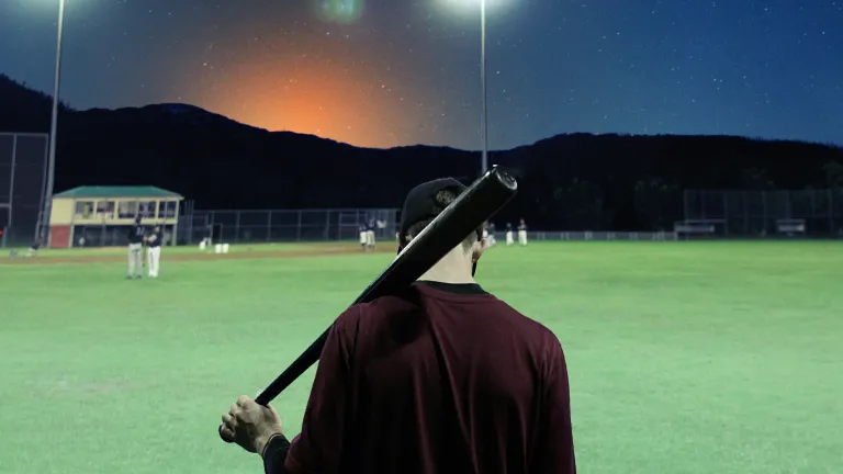 A young man holding a baseball bat on an empty ballfield at dusk.
