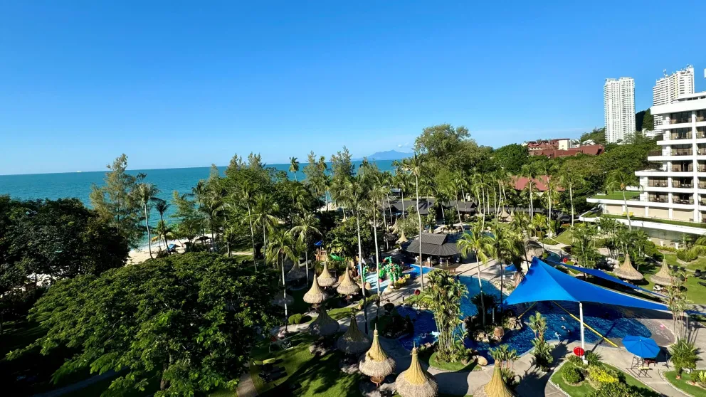 a seaside resort with blue and thatch umbrellas, a high rise building, and lots of green trees