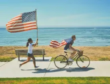 Man on bicycle and girl running behind with American flags.