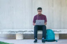 a man sitting ona bench and looking at a laptop with a backpack on the ground beside him