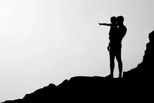 a silhouette of a father holding his son while standing on a rocky surface; the son is pointing into the distance
