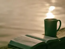 An open Bible and a ceramic mug sitting on a table with a pond in the background.