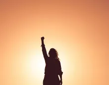 a woman's silhouette with a triumphant raised fist against a golden orange sky