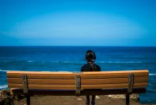 A woman sitting on a bench looking out on a large lake.