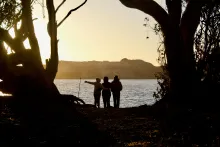 Three people watching the glow of the sun while they are standing by a lake.